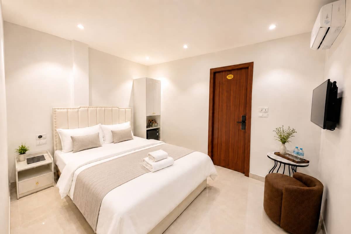 A modern hotel room with a large bed, bedside tables, a wardrobe, a small seating area, and a TV mounted on the wall.