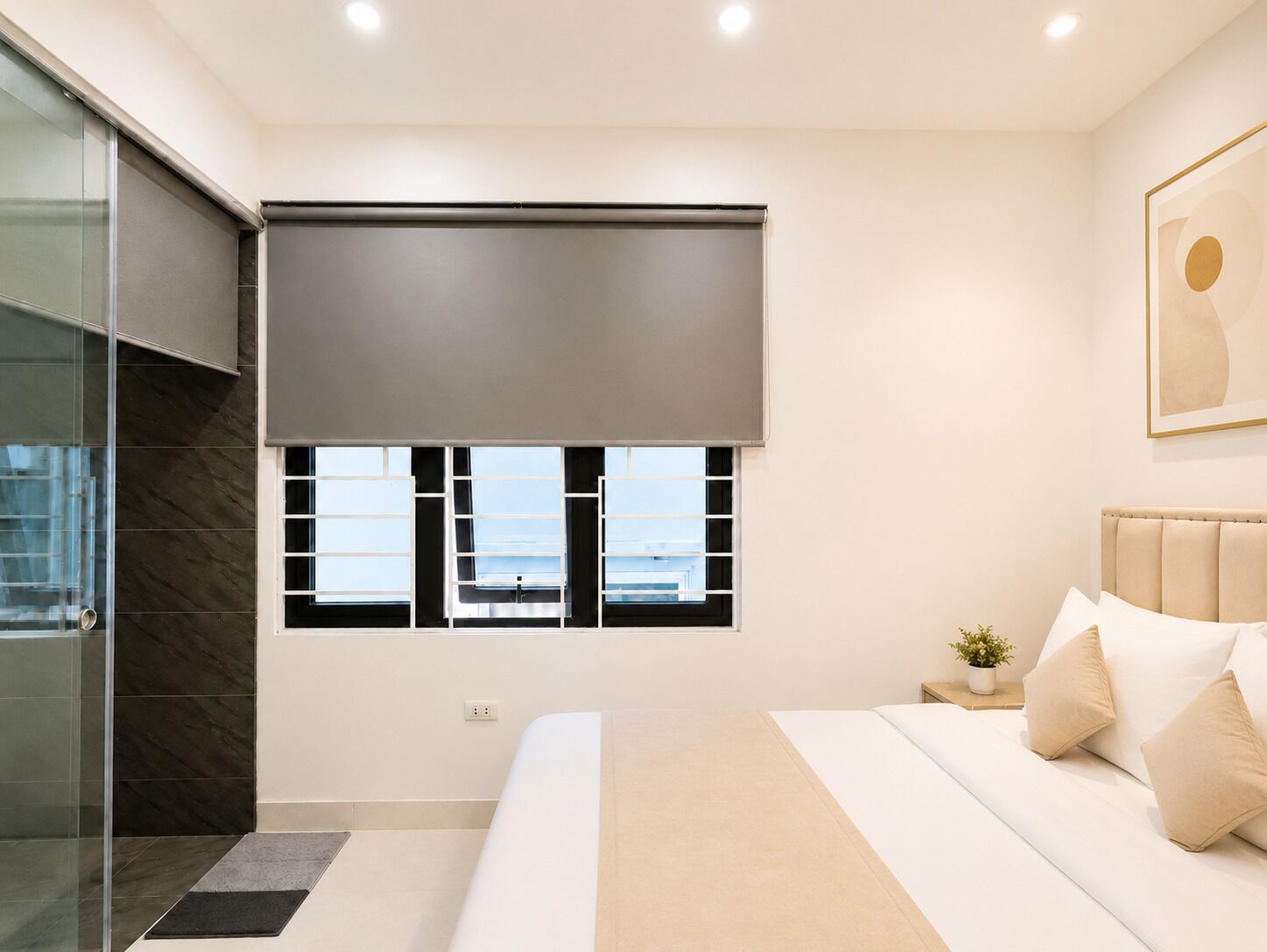 A modern bedroom with a large bed, a window with blinds, a bedside table with a plant, and a glass shower area.