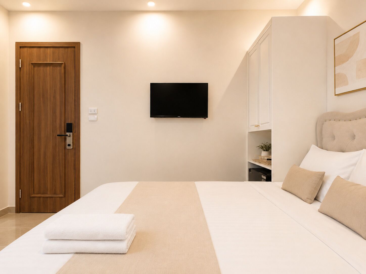 A hotel room with a wooden door, a flat-screen TV, a bed with white and beige bedding, and a headboard with storage.