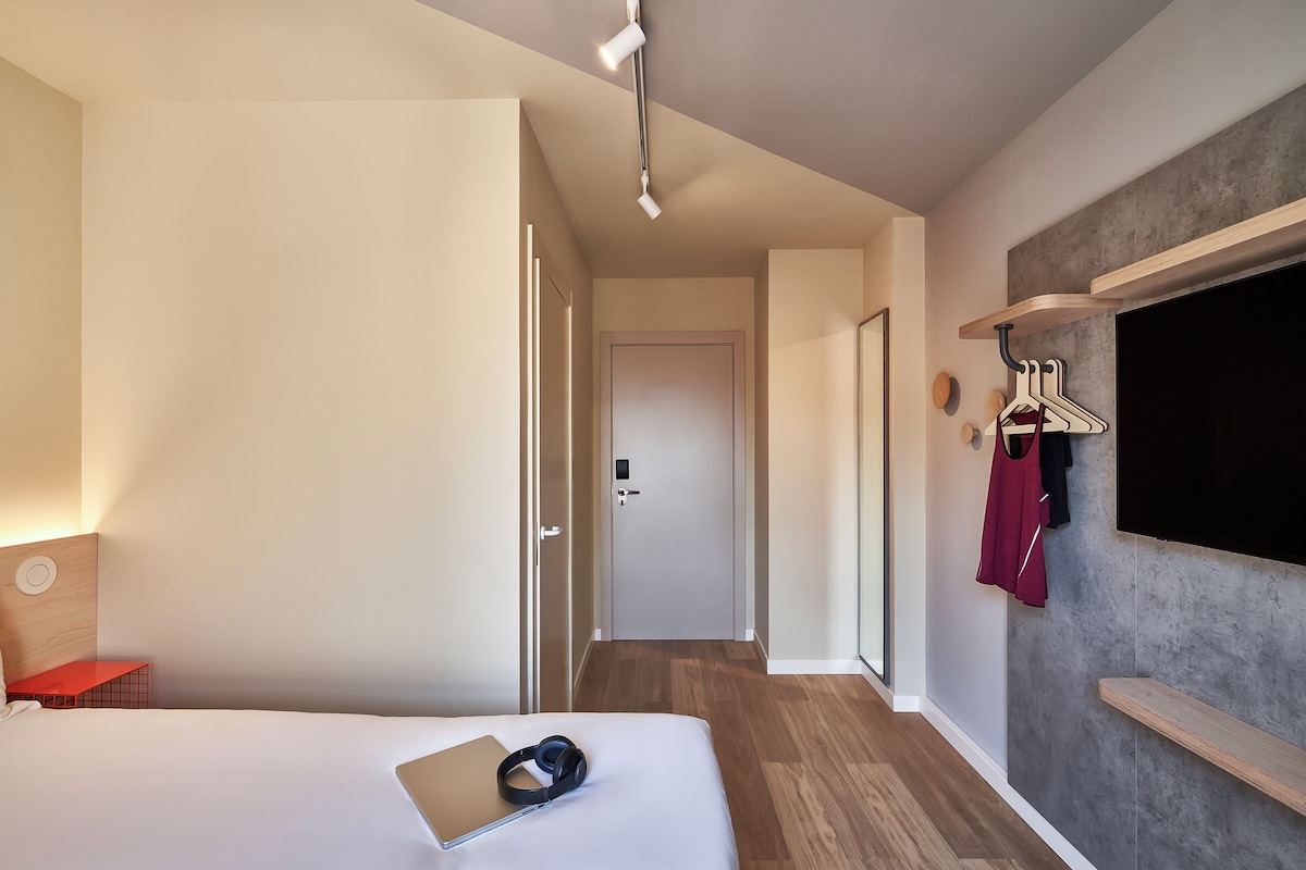 A hotel room with a bed, a desk, a TV mounted on the wall, and a hallway leading to another room.