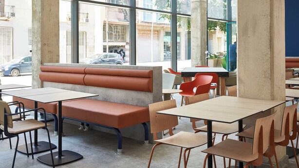 A modern cafe interior with high windows, a central bench, and multiple tables and chairs. - Ibis Budget Barcelona Center (Barcelona)