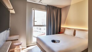 A hotel room with a bed, a desk, a chair, a window with curtains, and a view of buildings outside. - Ibis Budget Barcelona Center (Barcelona)