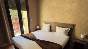 Ein Hotelzimmer mit einem großen Bett, einem Holz-Kopfbrett, zwei Nachttischen mit Lampen und Blick auf einen Balkon mit roten Blumen.