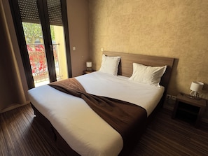 A hotel room with a large bed, wooden headboard, two bedside tables with lamps, and a view of a balcony with red flowers.