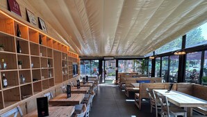 Ein Restaurantinneres mit Holztischen und -stühlen, einer Wand mit Regalen voller Flaschen und großen Fenstern.