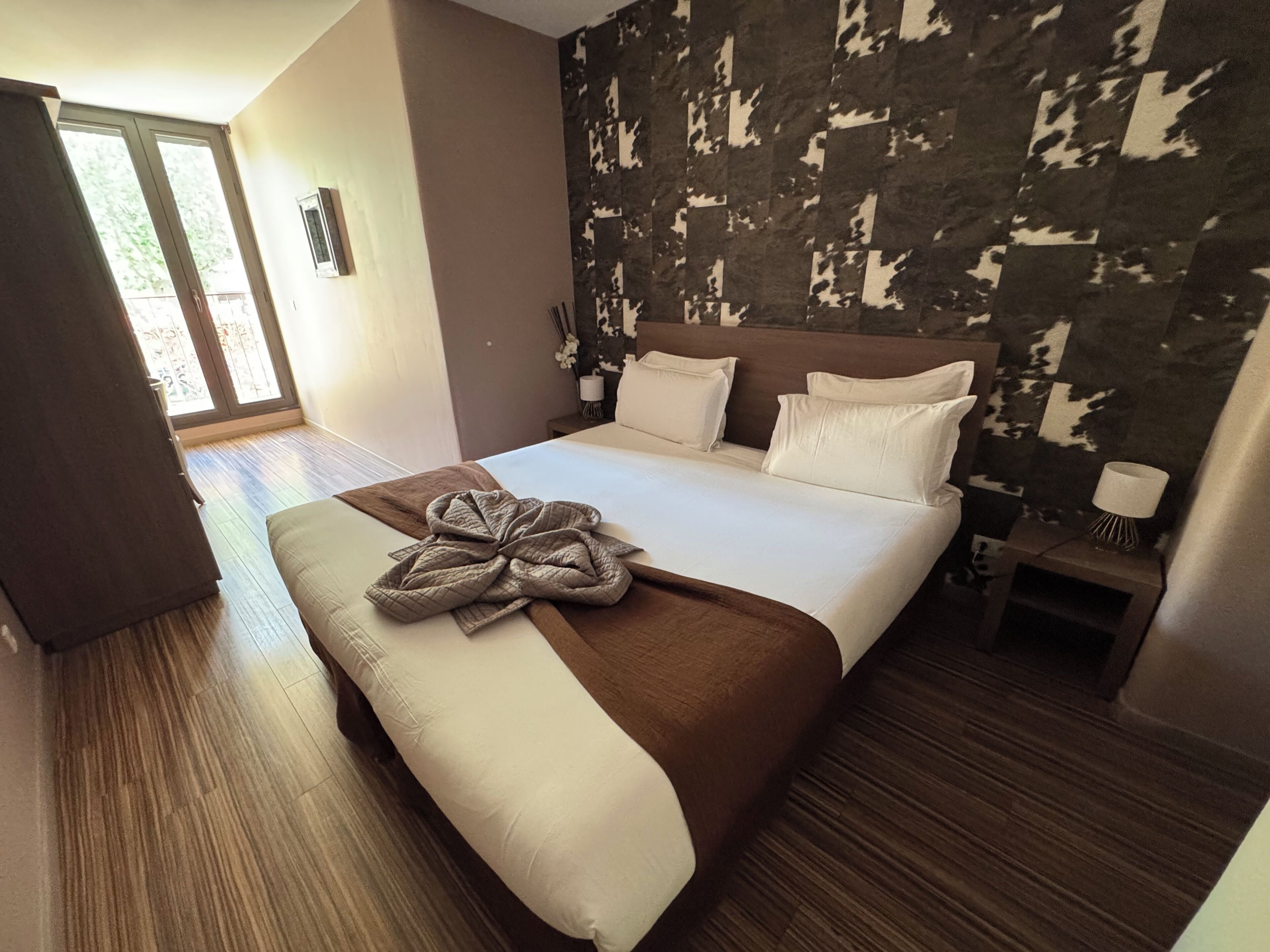 Habitación de hotel con una cama grande, suelo de madera, vista a través de un ventanal y pared con diseño.