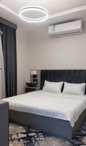 A modern hotel room with a bed, bedside table, lamp, air conditioning unit, and a circular light fixture.