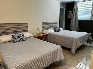 Two single beds with striped headboards, a bedside table with a lamp, and a window with curtains. - Cozy Downtown Isabela Private 2-bedroom Apt. near Park, Plaza & Beaches (Isabela)