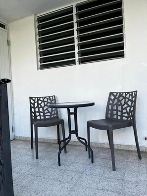 Un pequeño patio con dos sillas y una mesa, una ventana con persianas horizontales y una pared blanca.