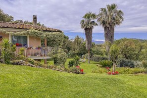 Ein Haus mit überdachter Terrasse, umgeben von einem gepflegten Garten mit Palmen und blühenden Pflanzen.