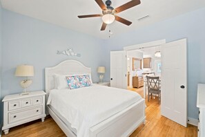 Une chambre à coucher avec un lit blanc, des tables de chevet, un ventilateur de plafond et une porte donnant sur une salle de bain.