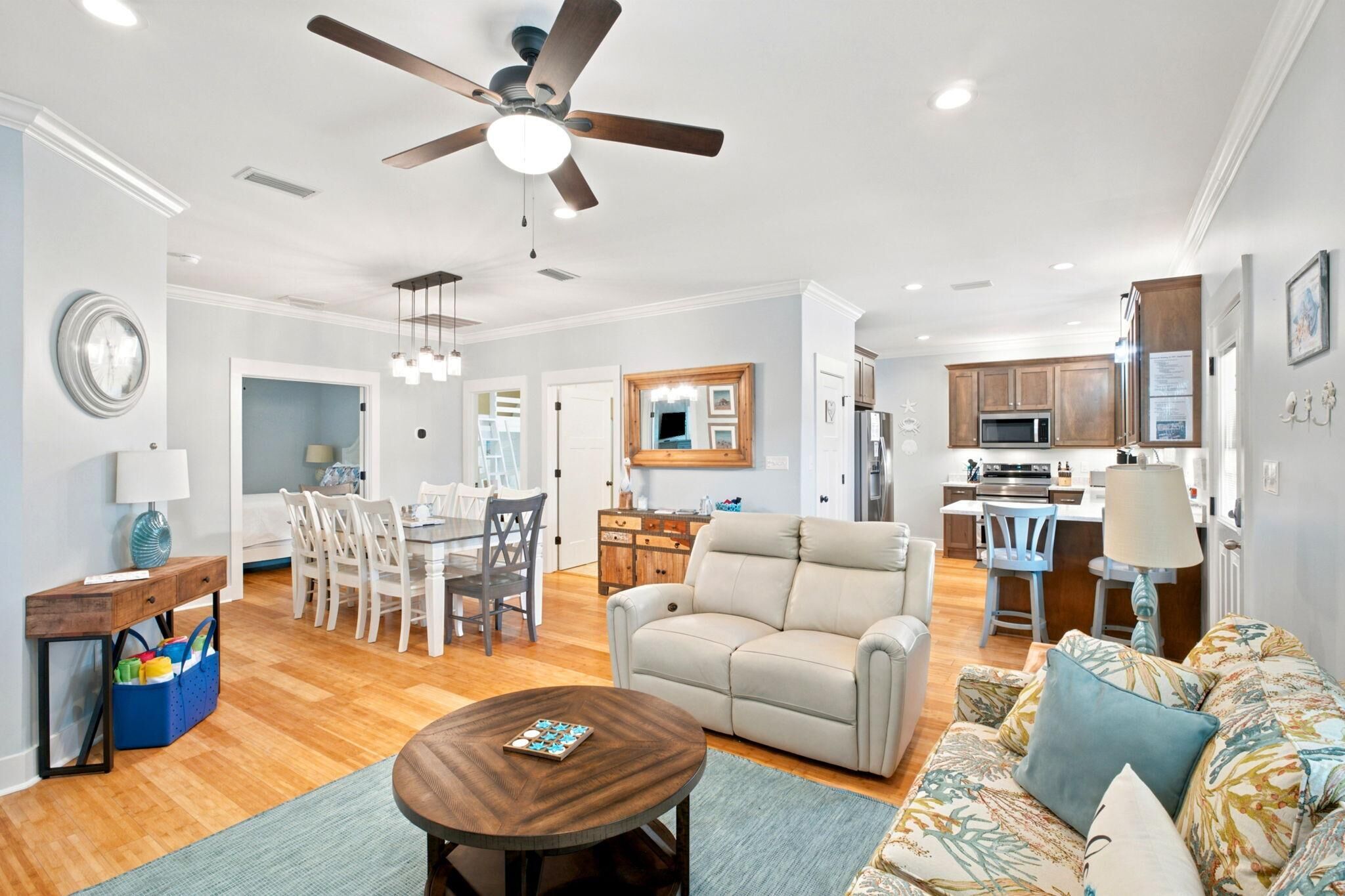 A modern living room with a sofa, a coffee table, a dining area with chairs, a kitchen with cabinets and appliances, and a ceiling fan.