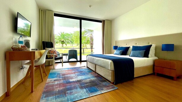 A hotel room with a bed, desk, chair, and a view of greenery outside.