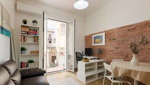 Een woonkamer met een bank, een wit bureau met een computer, een eettafel met stoelen, een boekenkast en een metselwerk muur.