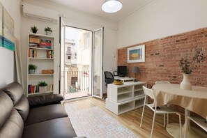 Ein Wohnzimmer mit einer Couch, einem weißen Schreibtisch mit Computer, einem Esstisch mit Stühlen, einem Bücherregal und einer Ziegelwand.