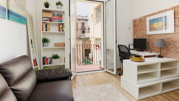Een moderne woonkamer met een leren bank, een witte boekenkast, een bureau met een computer, en uitzicht op een bakstenen gebouw door een open deur.