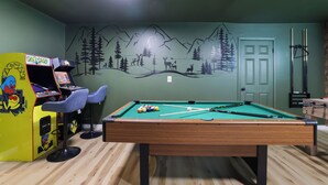 Uma mesa de bilhar com superfície de feltro verde, uma taça de bilhar e um conjunto de bolas. Há também uma máquina de jogos Pac-Man e dois bancos de bar. Um mural com paisagem de montanha está na parede.