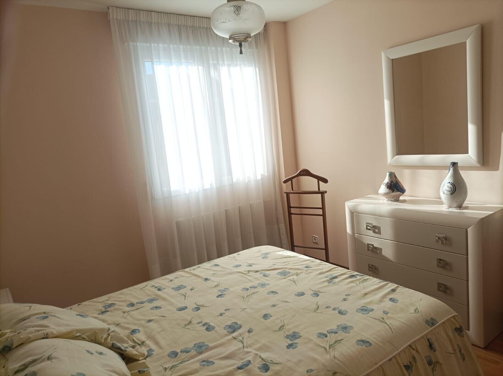 Een slaapkamer met een bed, een dressoir, een spiegel en een raam met dunne gordijnen.
