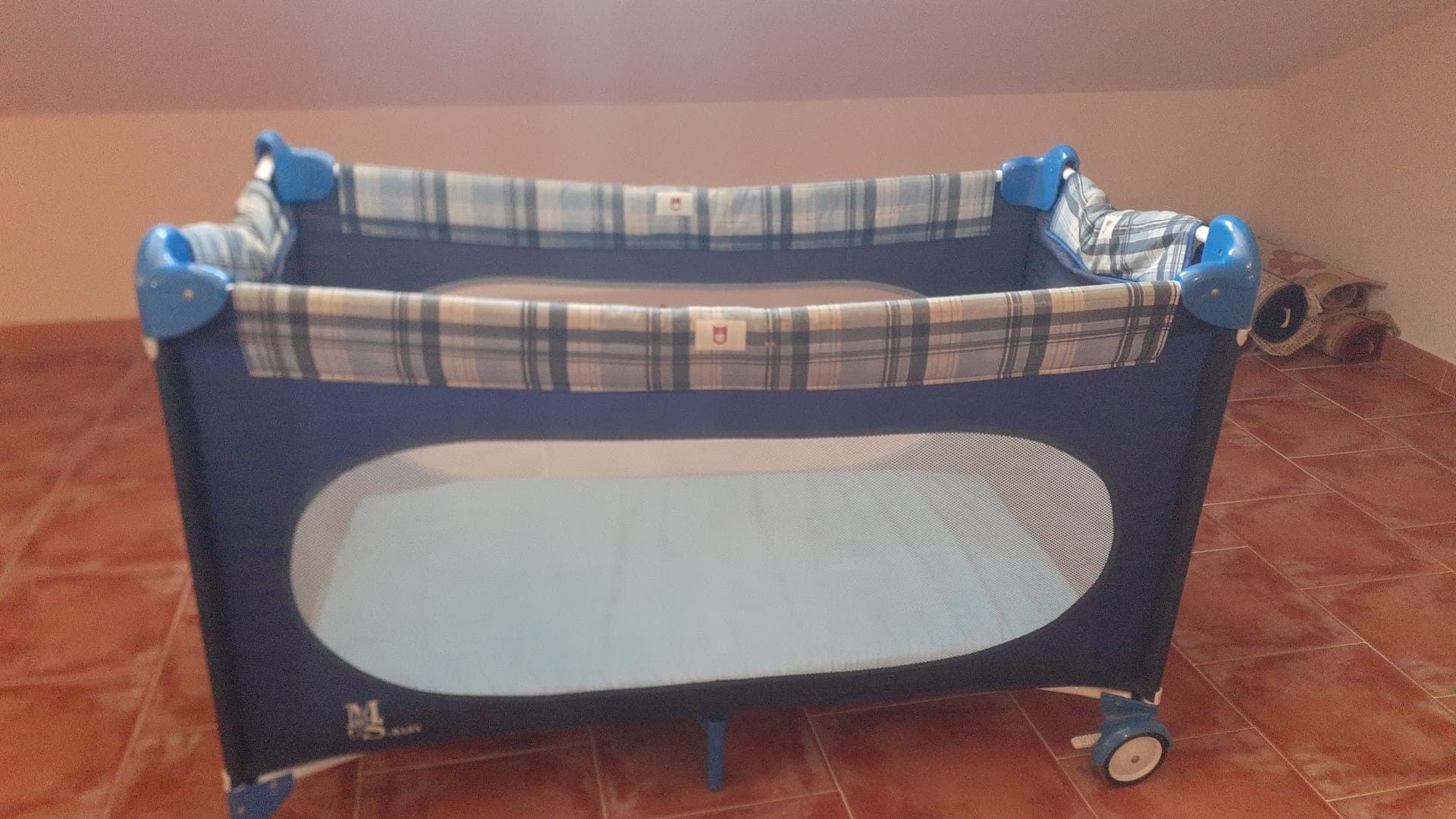 Una cuna para bebé con cubre sábana escocés, ruedas y una etiqueta.