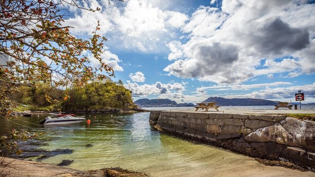 Une scène de bord de mer avec un bateau amarré près d’une jetée en pierre, un ciel dégagé et des montagnes lointaines.