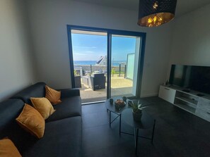 Una sala de estar moderna con un sofá, un televisor, una mesita con una planta y un ventanal corredero que da a un espacio exterior con vistas al mar.