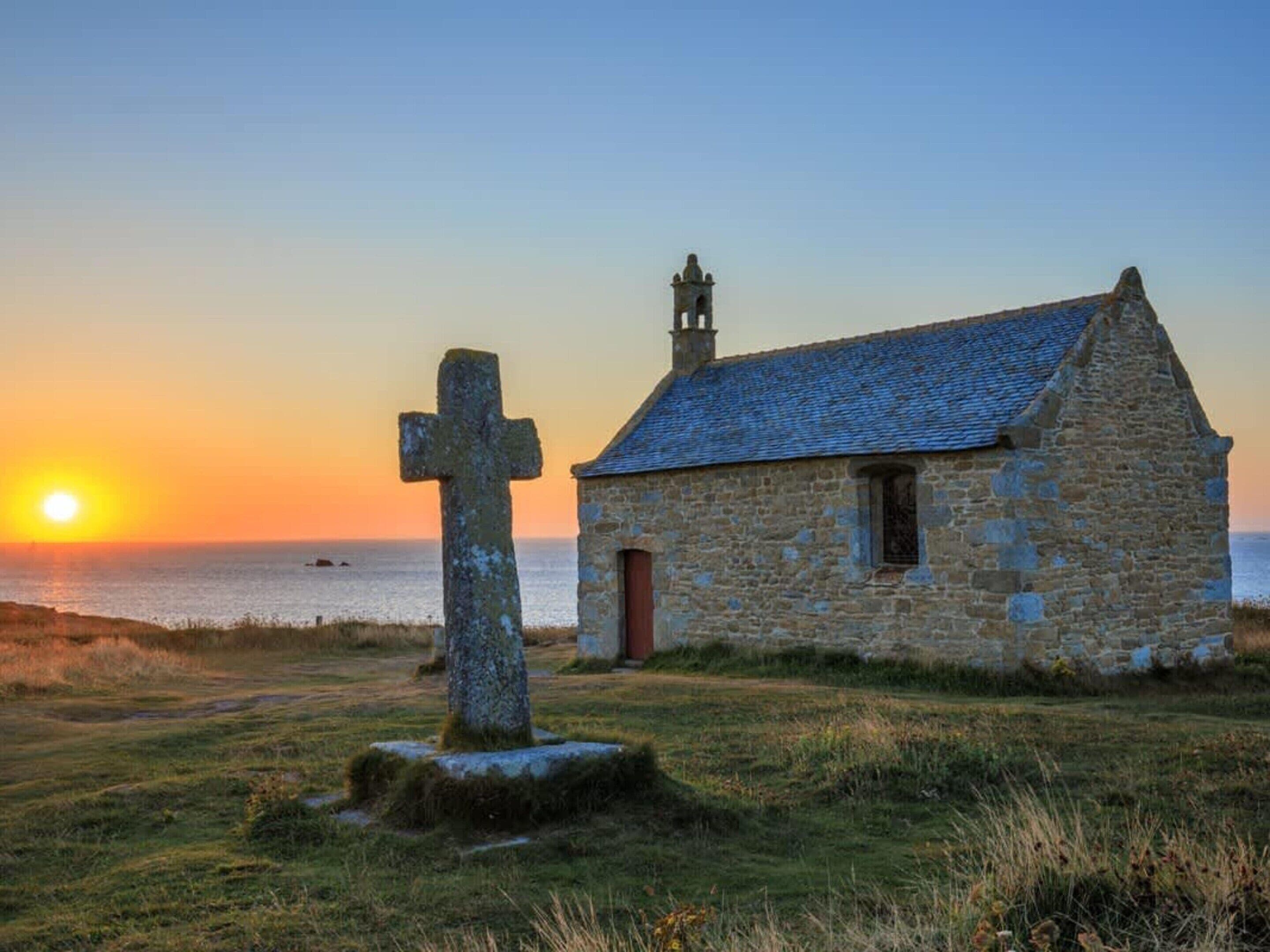 A stone church with a cross in front, a small tower, and a sunset over the sea.