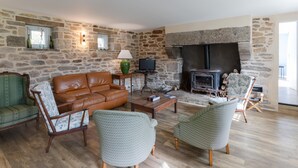 A stone-walled living room with a fireplace, leather sofa, patterned chairs, and a television. - Holiday Home by Lampaul Beach With Garden (Lampaul-Ploudalmézeau)