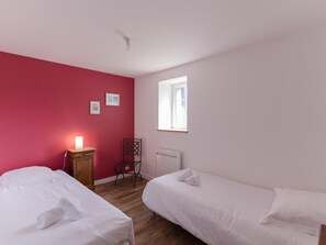 Ein Zimmer mit zwei Betten, einem Sessel, einem Schreibtisch mit Lampe, einem Fenster und einer Heizung.
