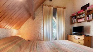 Ein Schlafzimmer mit schrägem Dach, einem Bett mit gestreifter Bettdecke, einer Holzkommode mit Fernseher und einem Bücherregal.