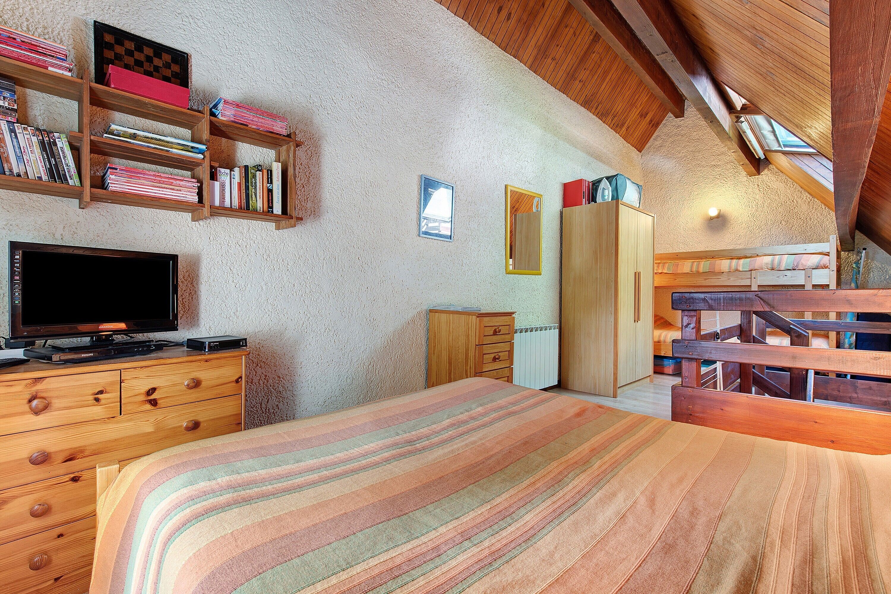 Un dormitorio con cama, cómoda de madera, televisor, estanterías y tragaluz.