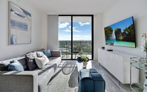 A modern living room with a sofa, a coffee table, a TV, and a balcony with a city view. - Modern 2BR Residence with open views & Balcony (Doral)