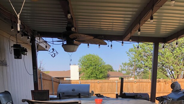Eine Terrasse im Freien mit einem Holztisch, Metallstühlen mit roten Kissen, einem Grill und einem Holzzaun im Hintergrund.