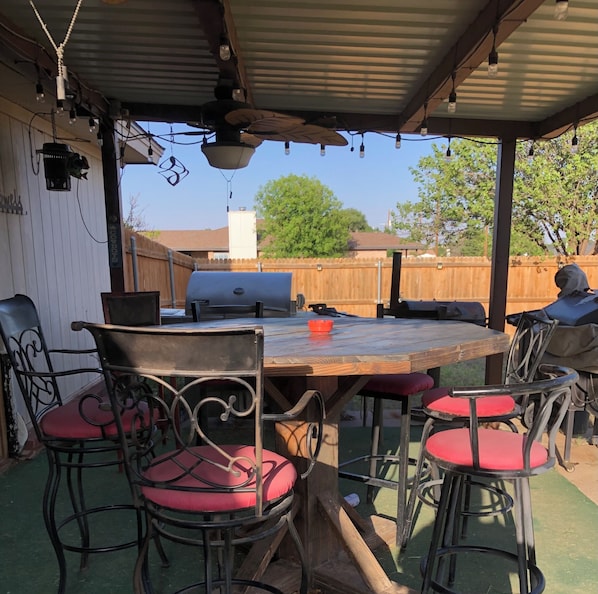 Eine Terrasse im Freien mit einem Holztisch, Metallstühlen mit roten Kissen, einem Grill und einem Holzzaun im Hintergrund.