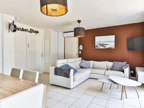 Ein modernes Wohnzimmer mit einem Ecksofa, einem runden Couchtisch, einem Esstisch mit Stühlen, einem Wand-Fernseher und einem Schild mit der Aufschrift 'Barber Shop'.