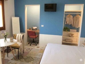 Ein Schlafzimmer mit einem Bett, einem Schreibtisch mit Laptop, einem Sessel, einem Fernseher und einem Kleiderschrank.