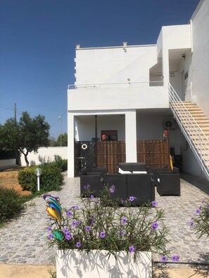 Un edificio moderno con un área de estar al aire libre cubierta, una escalera y un jardín con flores púrpuras.