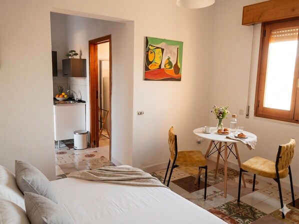 Un dormitorio con cama, una mesa de comedor con sillas, un cuadro en la pared y una ventana.