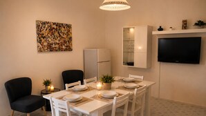 A dining area with a white table set for four, two chairs, a refrigerator, a cabinet with glass doors, and a flat-screen TV mounted on the wall. - Aqua Home Area – Core Five (Porto Cesareo)