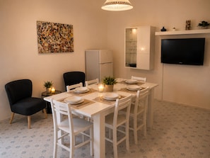 A dining area with a white table set for four, two chairs, a refrigerator, a cabinet with glass doors, and a flat-screen TV mounted on the wall. - Aqua Home Area – Core Five (Porto Cesareo)