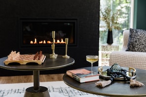 A cozy room with a fireplace, a chessboard, a book, a glass of wine, and a decorative vase on a round table. - Emerald Acres, an elevated Retreat on Five Private Acres (Bellingham)