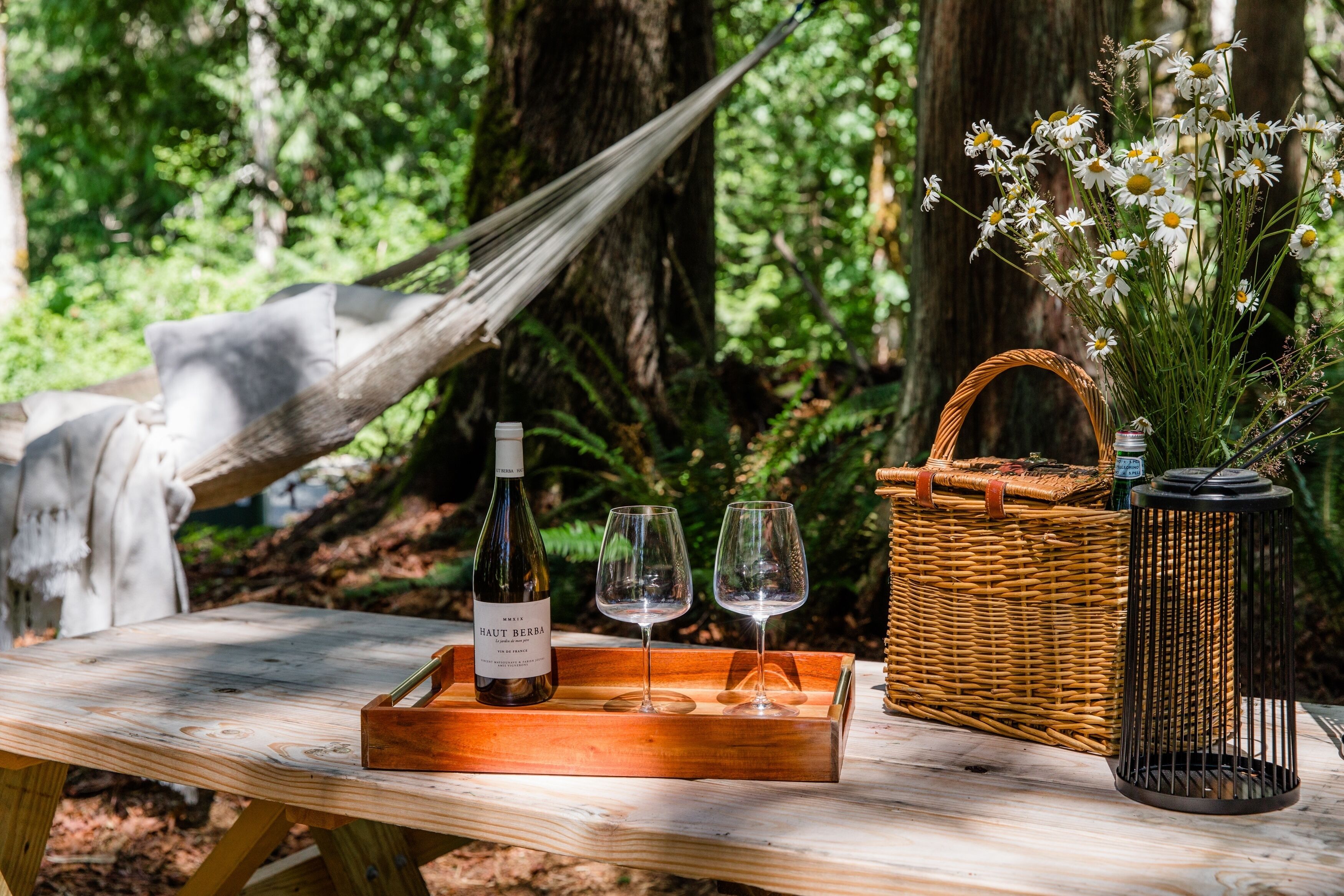 Ein Holztisch mit einer Weinflasche, zwei Gläsern, einem Korbkörbchen und einem Blumenstrauß in einer Waldlandschaft.