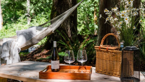 Ein Holztisch mit einer Weinflasche, zwei Gläsern, einem Korbkörbchen und einem Blumenstrauß in einer Waldlandschaft.