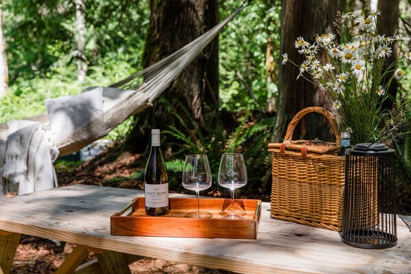 A wooden table with a wine bottle, two glasses, a wicker basket, and a vase of flowers in a forest setting. - Emerald Acres, an elevated Retreat on Five Private Acres (Bellingham)