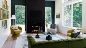 Ein modernes Wohnzimmer mit Kamin, einem grünen Sofa, Sesseln und großen Fenstern.