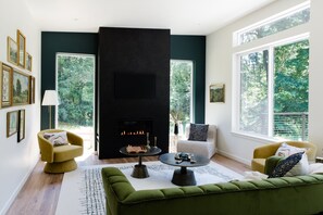 A modern living room with a fireplace, green sofa, armchairs, and large windows. - Emerald Acres, an elevated Retreat on Five Private Acres (Bellingham)