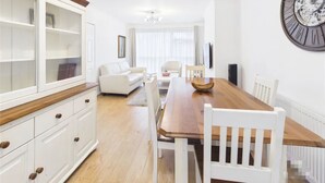 A modern living room with a dining area, white furniture, a large wooden dining table, and a wall-mounted clock. - 3 Bedroom Home with Private Garden in Newcastle (Whickham)