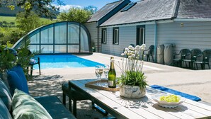 Ett bord vid poolkanten med en flaska, glas och en skål med vindruvor, omgivet av stolar och ett hus med en pool som har ett tak.