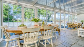 Ein Wintergarten mit einer großen Glasdoor, einem Holztisch, weißen Stühlen und Blick ins Freie.
