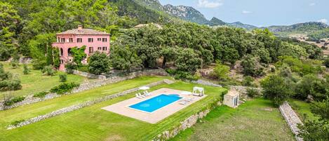A large pink villa with a swimming pool, surrounded by lush greenery and mountains.