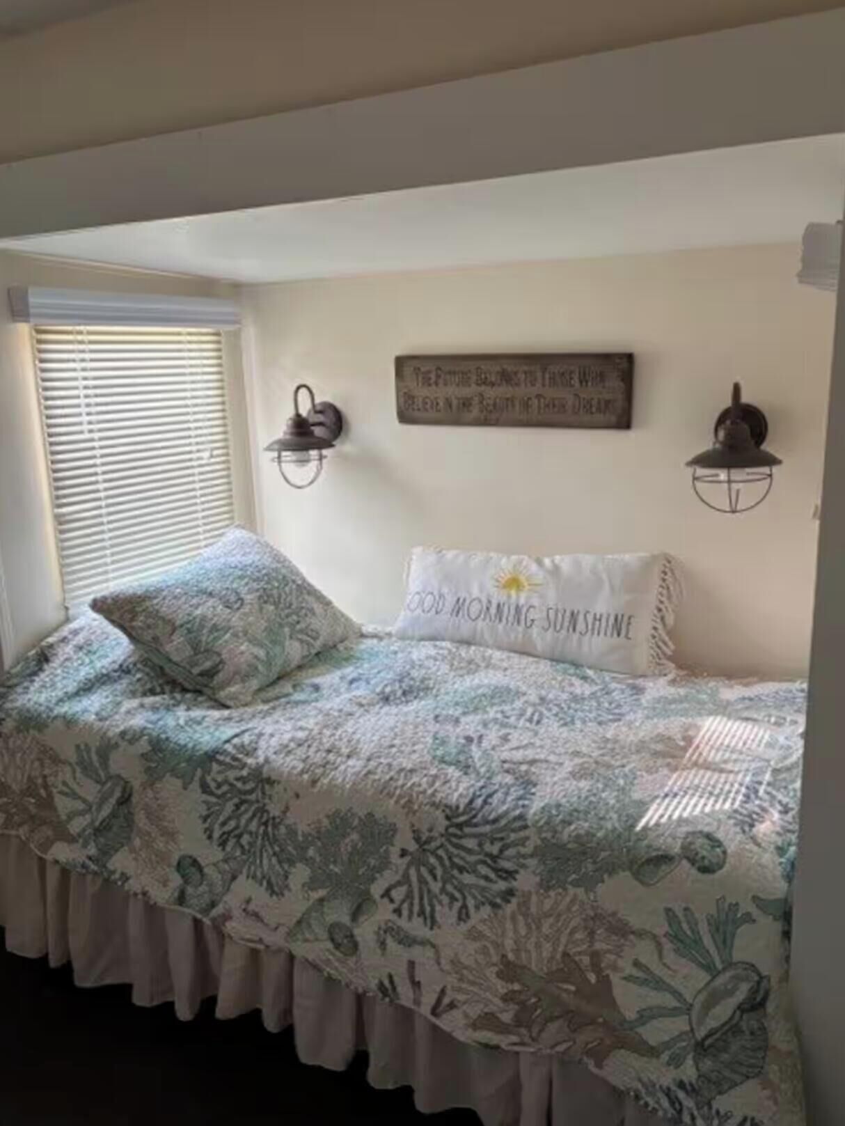 Ein Schlafzimmer mit Bett, Fenster mit Jalousien, einer Wandleuchte und einem motivierenden Zitat an der Wand.
